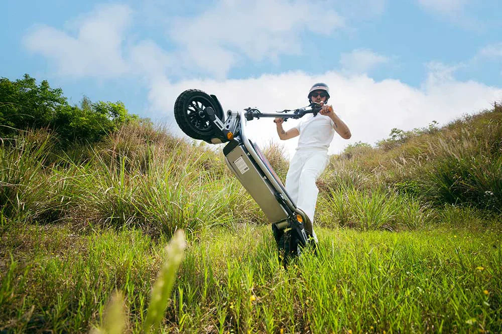 A young man riding a silver Kukirin A1 electric scooter across a green grassy field.